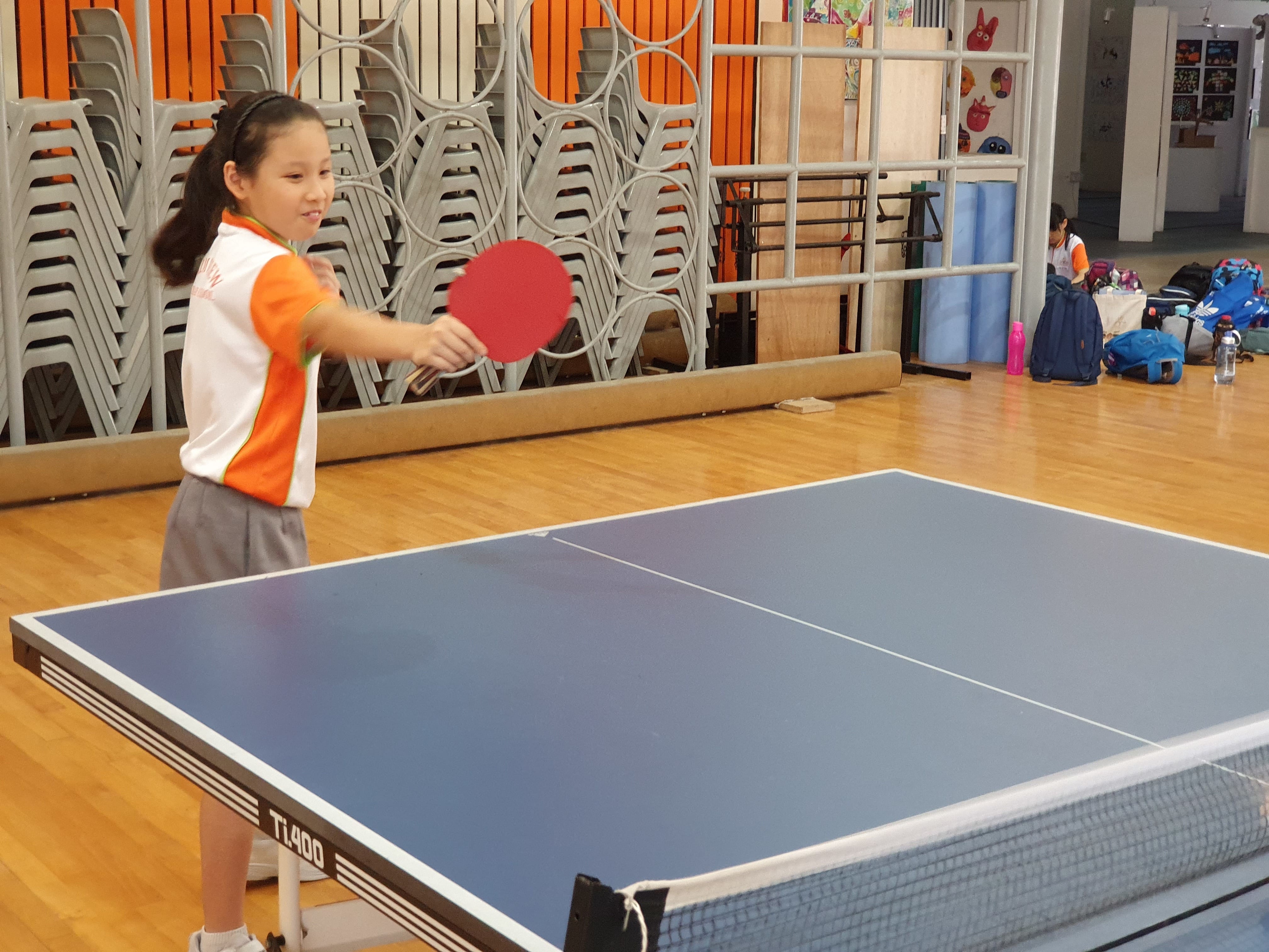 A young girl in a school uniform holds a red ping pong paddle beside a blue table tennis table indoors.